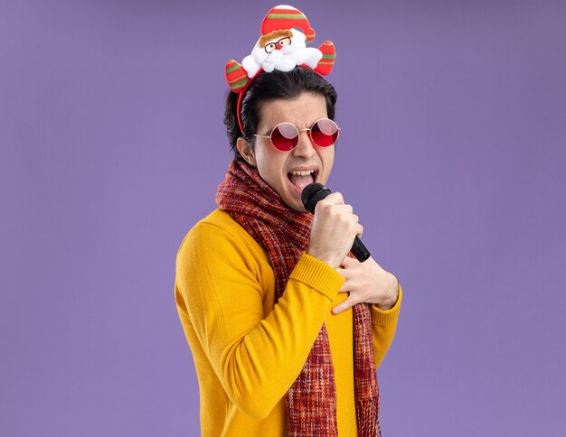 Joyful young man with warm scarf around neck in yellow turtleneck and glasses with funny rim on head holding microphone singing standing over purple background