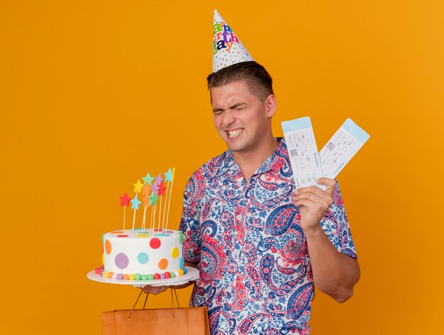 오렌지에 고립 된 선물 가방과 티켓으로 케이크를 들고 생일 모자를 쓰고 즐거운 젊은 파티 남자