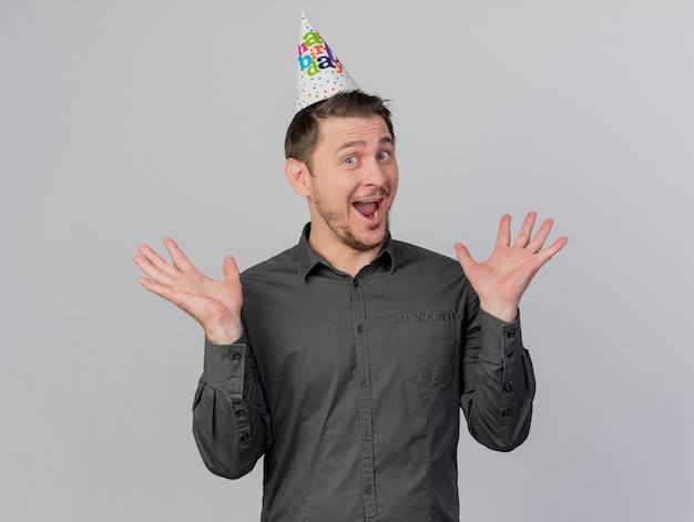 Joyful young party guy wearing birthday cap spreading hands isolated on white