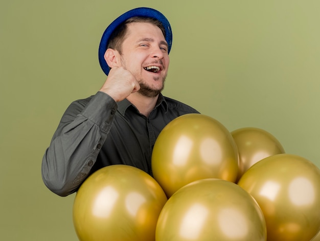 Joyful young party guy wearing black shirt and blue hat standing behind balloons showing yes gesture isolated on olive green