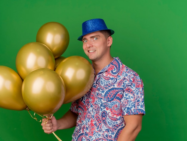 Joyful young party guy wearing blue hat holding balloons isolated on green