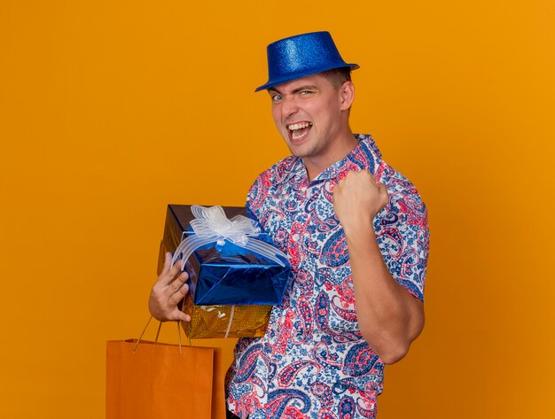 오렌지에 고립 예 제스처를 보여주는 가방 선물 상자를 들고 파란색 모자를 쓰고 즐거운 젊은 파티 남자