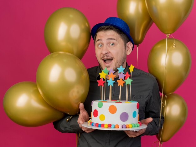 ピンクで隔離のケーキを保持している風船の間に立っている青い帽子をかぶってうれしそうな若いパーティーの男