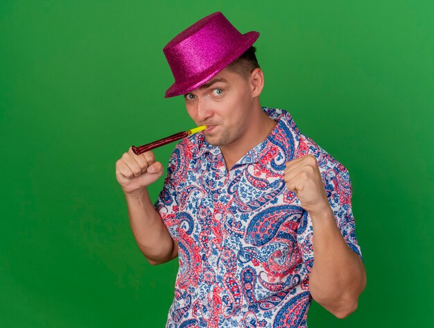 Joyful young party guy wearing pink hat blowing party blower showing yes gesture isolated on green background