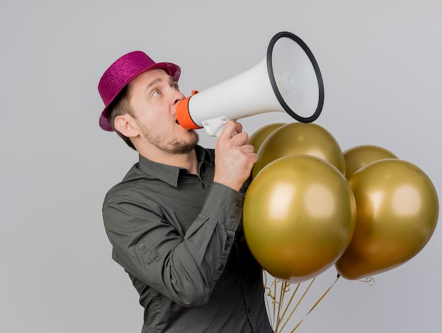 Joyful young party guy  wearing pink hat holding balloons and speaks on loudspeaker isolated on white