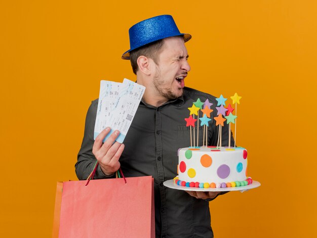 오렌지에 고립 된 티켓 선물 가방과 케이크를 들고 파란색 모자를 쓰고 눈을 감고 즐거운 젊은 파티 남자