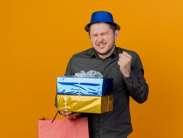 Радостный молодой тусовщик с закрытыми глазами в синей шляпе держит подарочные коробки и сумку, показывая жест `` да '', изолированный на оранжевом