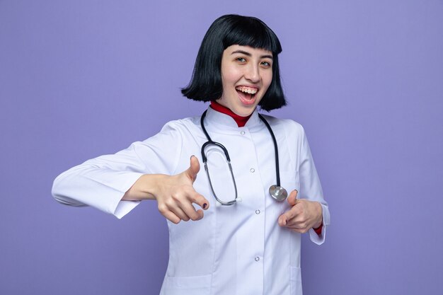 Joyful young pretty caucasian girl in doctor uniform with stethoscope pointing at front with two hands