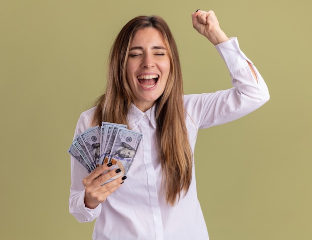 Foto gratuita la giovane ragazza abbastanza caucasica allegra tiene i soldi e tiene il pugno su isolato sulla parete verde oliva con lo spazio della copia