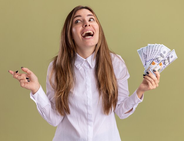 Joyful young pretty caucasian girl holds money looking and pointing at side on olive green 