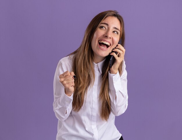 Радостная молодая симпатичная кавказская девушка держит кулак и разговаривает по телефону, глядя в сторону на фиолетовом
