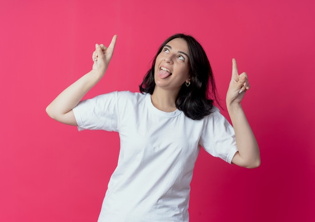 Joyful young pretty caucasian girl showing tongue looking and pointing up isolated on crimson background