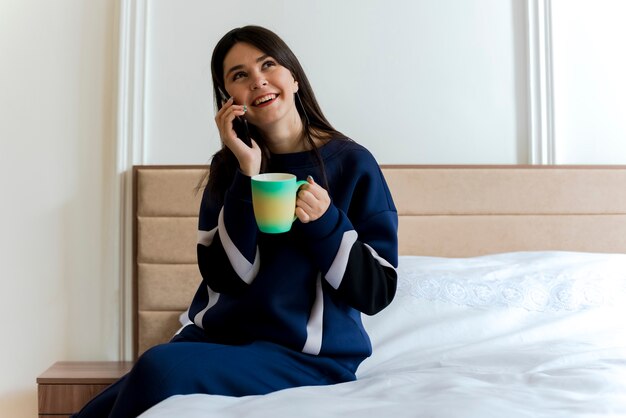 Радостная молодая симпатичная кавказская девушка сидит на кровати в спальне, держа чашку, глядя в сторону и разговаривает по телефону