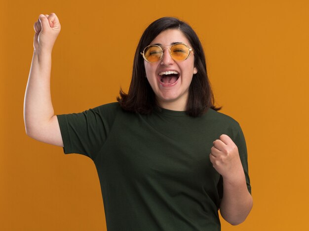 Joyful young pretty caucasian girl in sun glasses standing with raised fists isolated on orange wall with copy space