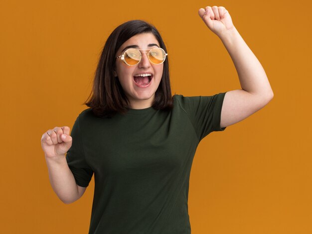 Joyful young pretty caucasian girl in sun glasses stands with raised fists up