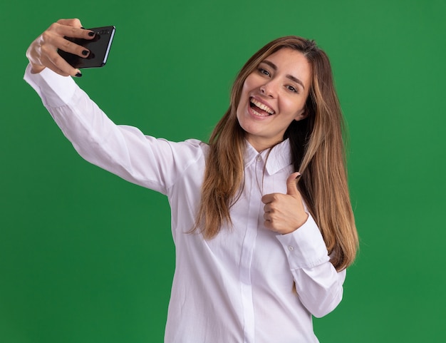 La giovane ragazza abbastanza caucasica allegra alza i pollici e tiene il telefono isolato sulla parete verde con lo spazio della copia