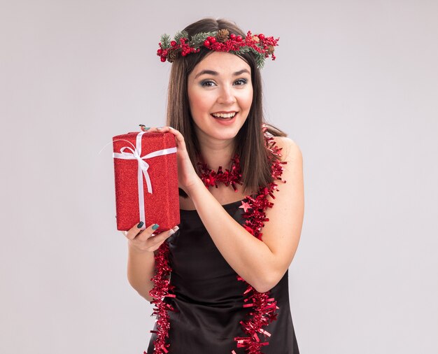 Радостная молодая симпатичная кавказская девушка в рождественском венке и гирлянде из мишуры на шее, держащая подарочный пакет, смотрит в камеру, изолированную на белом фоне с копией пространства