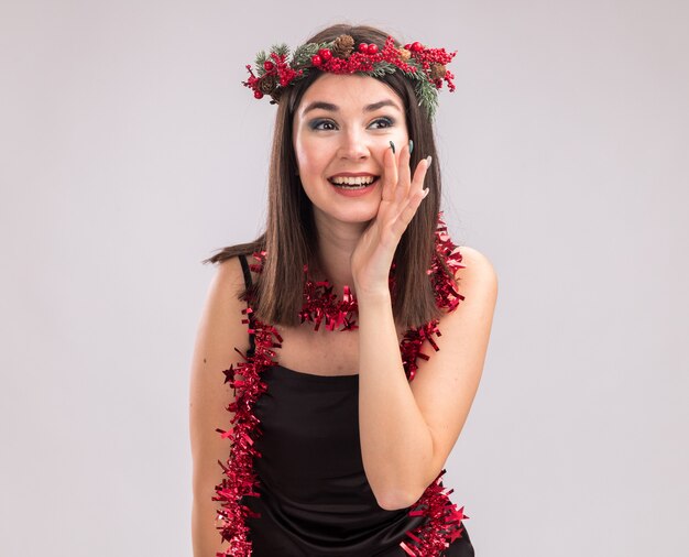 クリスマスの頭の花輪と首の周りに見掛け倒しの花輪を身に着けているうれしそうな若いかわいい白人の女の子は、コピースペースで白い背景に分離された側のささやきを見て