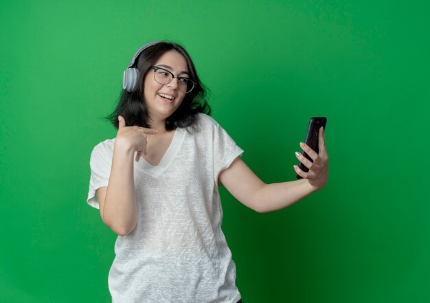 Joyful young pretty caucasian girl wearing glasses and headphones holding and looking at mobile phone and pointing at herself