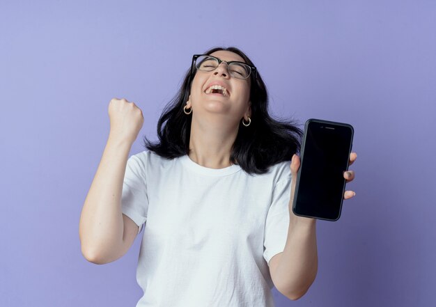 Joyful young pretty caucasian girl wearing glasses showing mobile phone and doing yes gesture with closed eyes isolated on purple background