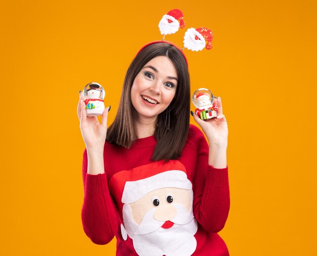 Joyful young pretty caucasian girl wearing santa claus headband and sweater holding snowman and santa claus figurines looking at camera isolated on orange background with copy space