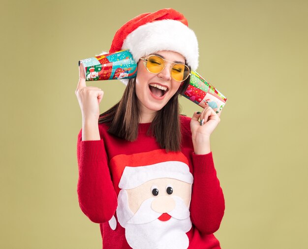 Joyful young pretty caucasian girl wearing santa claus sweater and hat with glasses holding plastic christmas cups next to ears listening to conversations with closed eyes