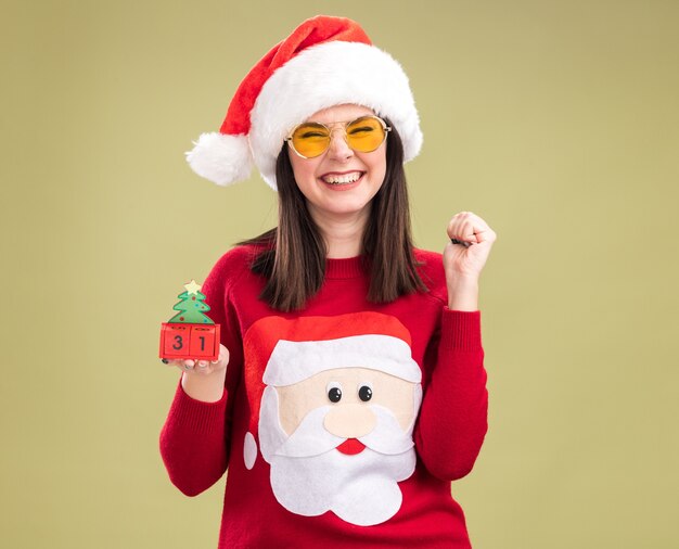 サンタクロースのセーターとヘッドバンドを身に着けているうれしそうな若いかわいい白人の女の子は、オリーブグリーンの背景に分離されたはいジェスチャーをしているカメラを見て日付とクリスマスツリーのおもちゃを保持しているメガネ