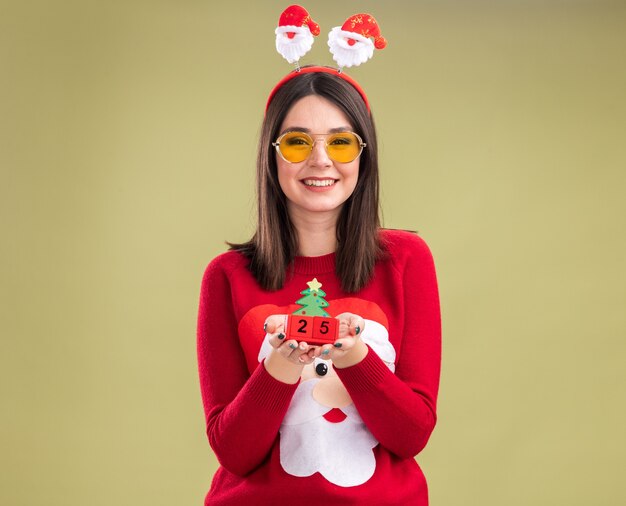 Joyful young pretty caucasian girl wearing santa claus sweater and headband with glasses holding christmas tree toy with date 