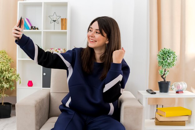 Joyful young pretty caucasian woman sitting on armchair in designed living room clenching fist and taking selfie