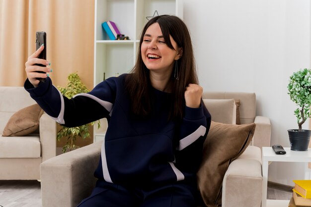 Радостная молодая симпатичная кавказская женщина, сидящая на кресле в дизайнерской гостиной, держит и смотрит на мобильный телефон и делает жест "да"