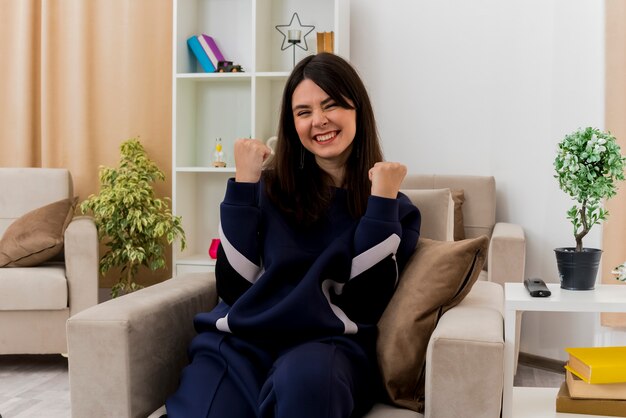 Радостная молодая симпатичная кавказская женщина, сидящая на кресле в дизайнерской гостиной, смотрит, делает жест да