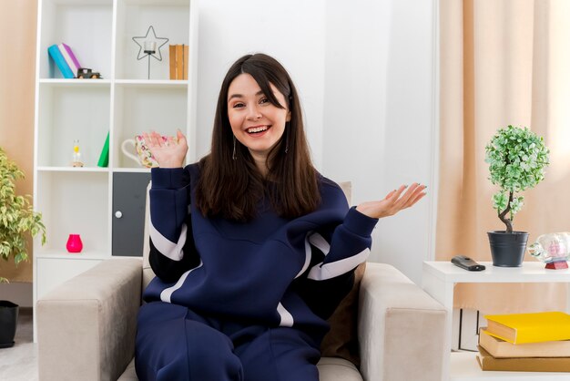 Joyful young pretty caucasian woman sitting on armchair in designed living room showing empty hands looking