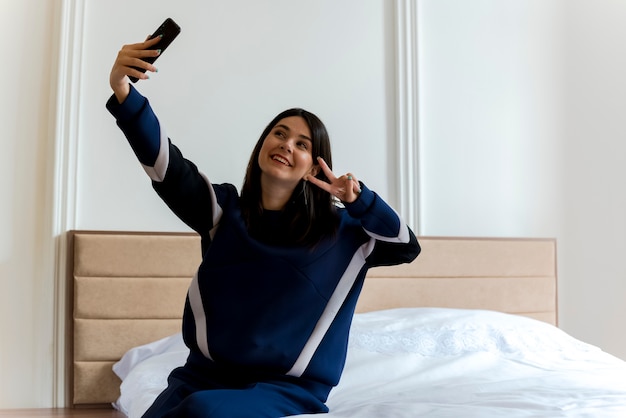 無料写真 ピースサインをして、selfieを取る寝室のベッドに座ってうれしそうな若いかなり白人女性