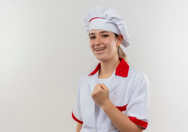 Joyful young pretty cook in chef uniform with dental braces clenching fist isolated on white wall with copy space