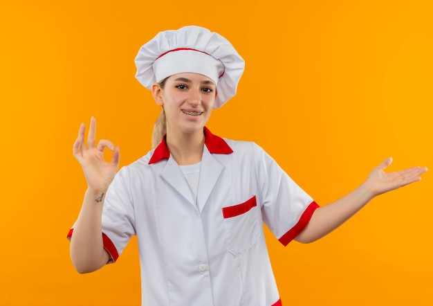 Gioioso giovane cuoco grazioso in uniforme da chef con parentesi graffe dentali facendo segno ok e mostrando la mano vuota isolata sullo spazio arancione