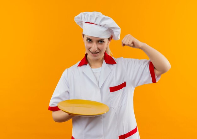 Joyful young pretty cook in chef uniform with dental braces holding empty plate and raising fist isolated on orange space