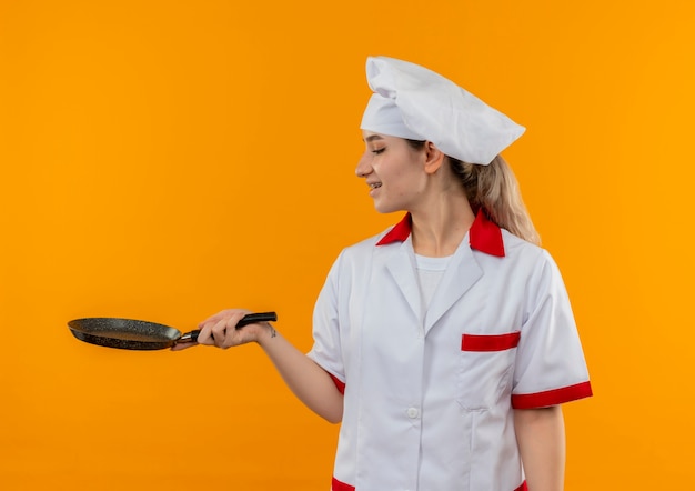 Joyful young pretty cook in chef uniform with dental braces holding and looking at frying pan isolated on orange space