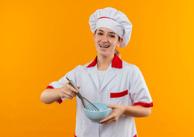 Радостный молодой симпатичный повар в униформе шеф-повара с зубными скобами держит венчик и миску, изолированные на оранжевом пространстве