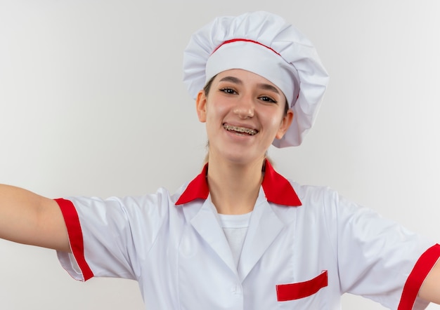 Радостный молодой симпатичный повар в униформе шеф-повара с брекетами смотрит с распростертыми объятиями, изолированными на белом пространстве