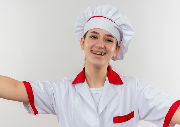 Joyful young pretty cook in chef uniform with dental braces looking  with open arms isolated on white space