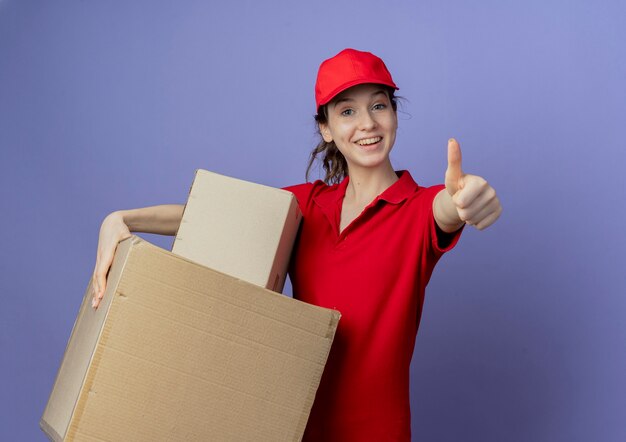 Joyful young pretty delivery girl wearing red uniform and cap holding carton boxes and showing thumb up at camera isolated on purple background with copy space