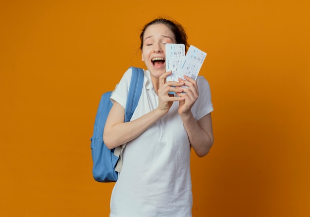복사 공간 오렌지 배경에 고립 된 닫힌 된 눈으로 비행기 티켓을 들고 다시 가방을 입고 즐거운 젊은 예쁜 여자 학생