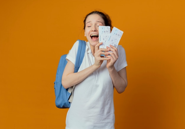 Joyful young pretty female student wearing back bag holding airplane tickets with closed eyes isolated on orange background with copy space