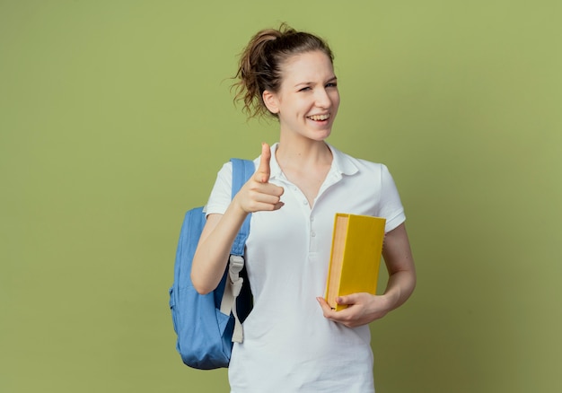 Радостная молодая симпатичная студентка в задней сумке держит книгу и указывает на камеру, изолированную на зеленом фоне с копией пространства