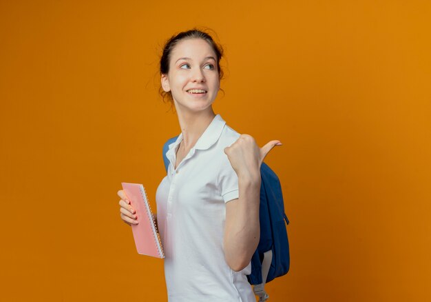 Joyful young pretty female student wearing back bag standing in profile view holding note pad looking and pointing behind isolated on orange background with copy space