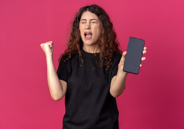 Joyful young pretty girl holding mobile phone doing yes gesture with closed eyes 