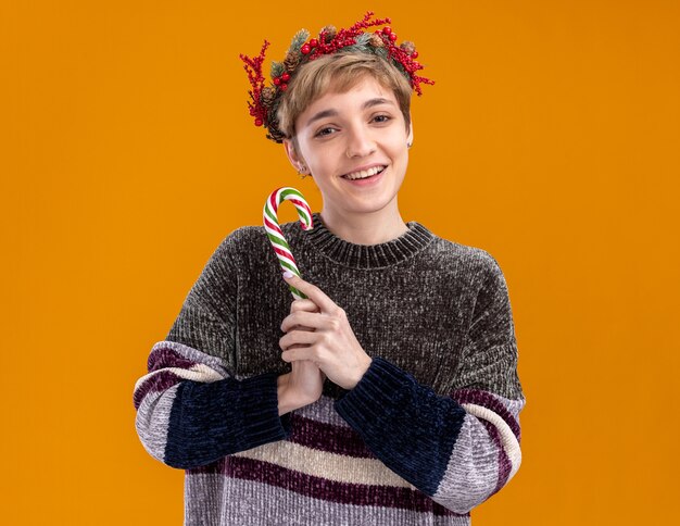 Joyful young pretty girl wearing christmas head wreath holding christmas sweet cane  isolated on orange wall