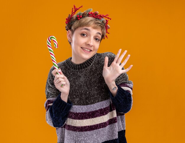 Joyful young pretty girl wearing christmas head wreath holding christmas sweet cane looking at camera showing empty hand isolated on orange background