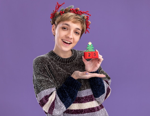 Радостная молодая красивая девушка в рождественском венке держит елочную игрушку с датой, указывающей на нее, изолированной на фиолетовой стене с копией пространства