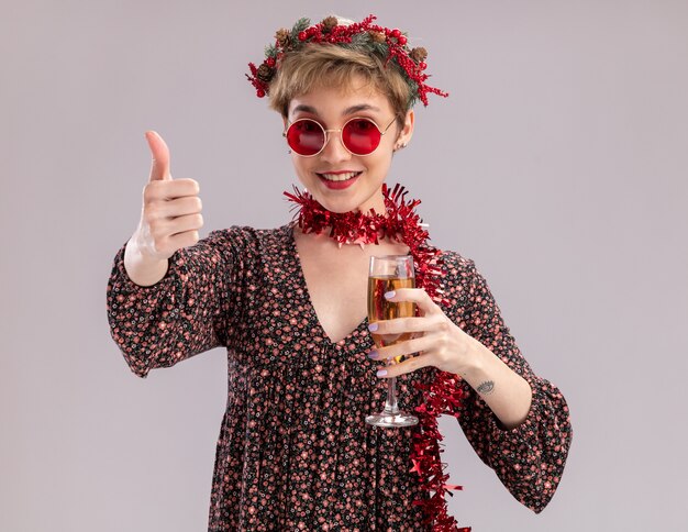 Joyful young pretty girl wearing christmas head wreath and tinsel garland around neck with glasses holding glass of champagne looking at camera showing thumb up isolated on white background